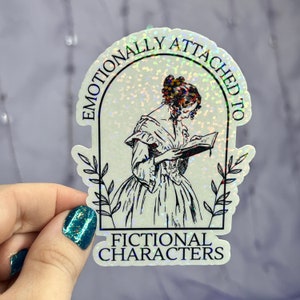 May include: A holographic sticker with a glittery background featuring a woman reading a book in a window frame. The text reads "Emotionally Attached to Fictional Characters".