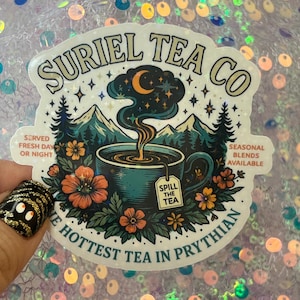 May include: A holographic sticker featuring the text "SURIEL TEA CO" with a teal teacup design. The design includes mountains, flowers, and the text "THE HOTTEST TEA IN PRYTHIAN". Additional text includes "SERVED FRESH DAY OR NIGHT" and "SEASONAL BLENDS AVAILABLE".