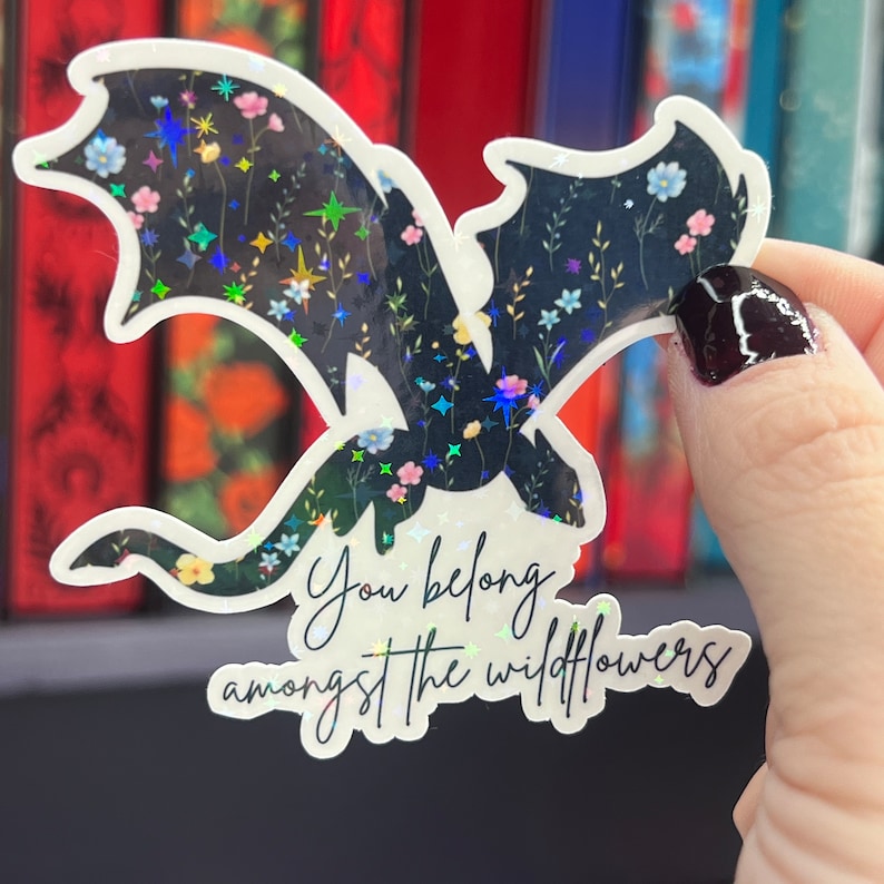 Op de afbeelding: Een zwart-witte sticker met een draken silhouet en een bloemenpatroon. De sticker heeft iriserende glitter en de tekst "You belong amongst the wildflowers".