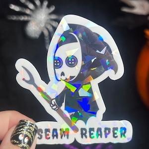 May include: A holographic sticker featuring a cartoon Grim Reaper holding a seam ripper. The text "SEAM REAPER" is printed below the image.