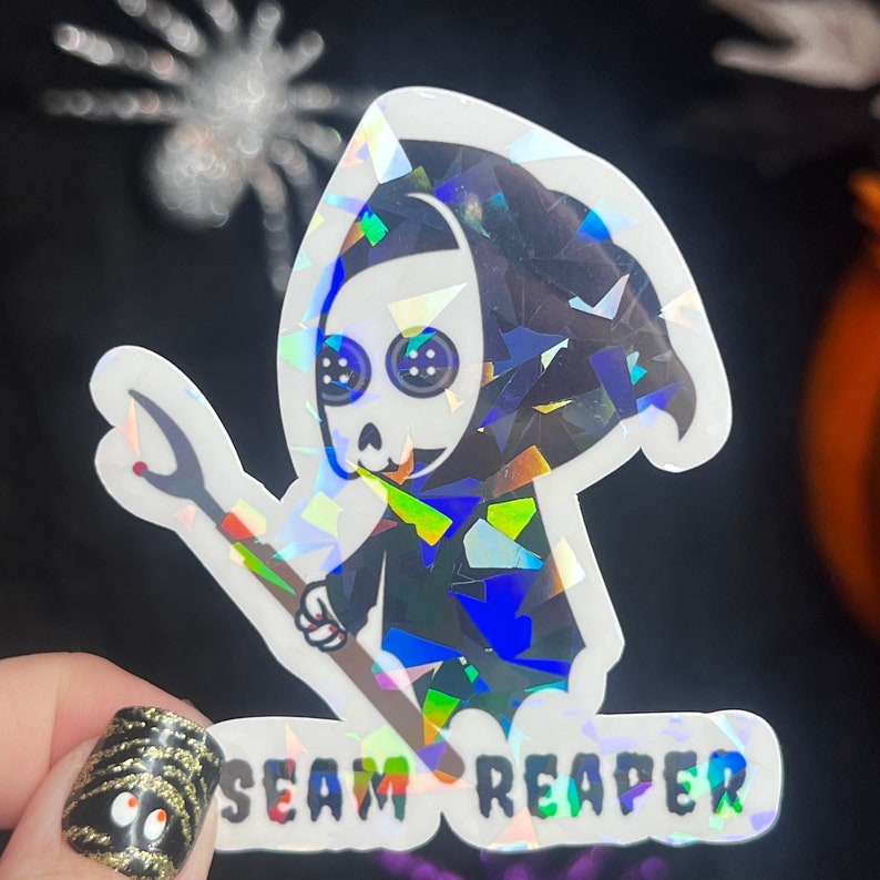 May include: A holographic sticker featuring a cartoon Grim Reaper holding a seam ripper. The text "SEAM REAPER" is printed below the image.