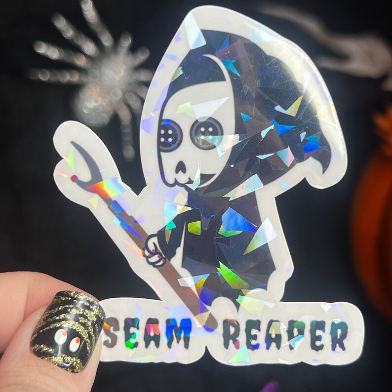 May include: A holographic sticker featuring a cartoon Grim Reaper holding a needle and thread. The sticker has the text "SEAM REAPER" at the bottom.