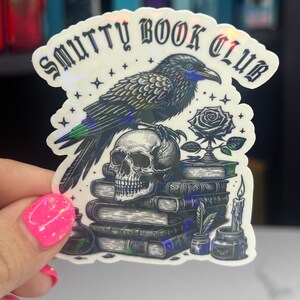 May include: A black and white sticker with a raven perched on a skull, a rose, and a stack of books. The sticker says "Smutty Book Club".