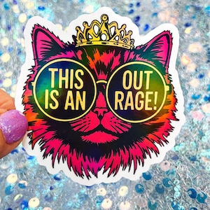 May include: A die-cut sticker featuring a vibrant, multi-colored cat head wearing a gold crown and round sunglasses. The lenses of the glasses display the text THIS IS AN OUT RAGE! in gold lettering against a dark, iridescent background.