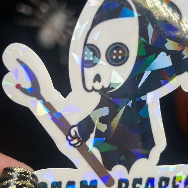 May include: A holographic sticker featuring a cartoon skeleton wearing a black hooded robe and holding a stick. The skeleton has large, round eyes with black buttons for pupils. The sticker has a glittery, iridescent finish.