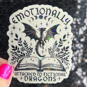 May include: A holographic sticker with a black and white illustration of a dragon, a book, and floral elements. The text reads "Emotionally Attached to Fictional Dragons".