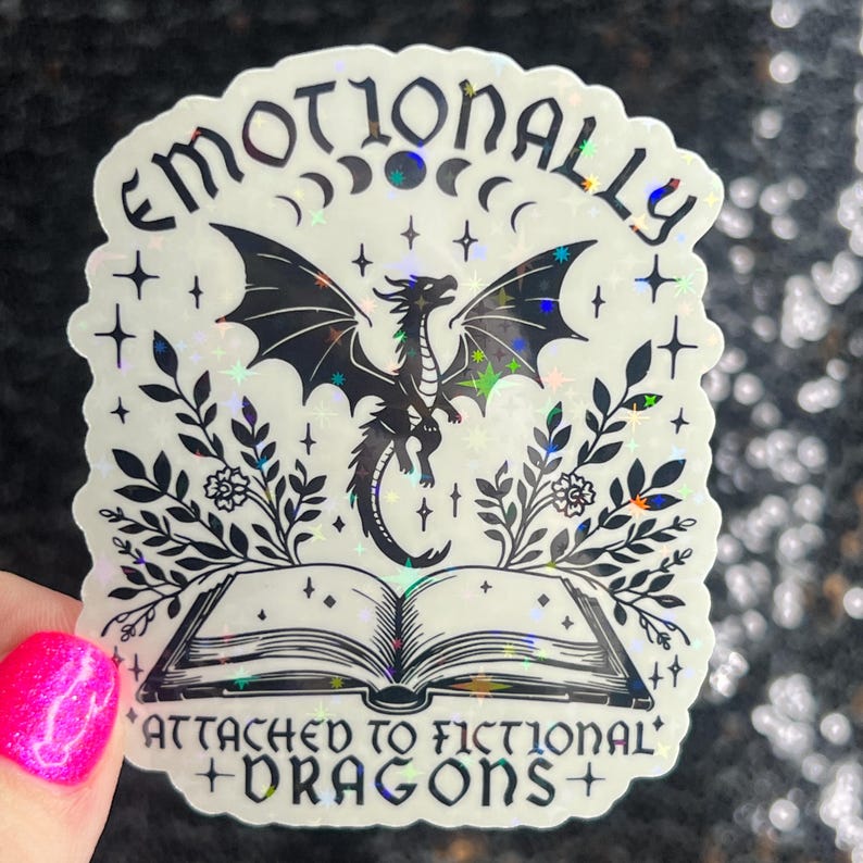 May include: A holographic sticker with a black and white illustration of a dragon, a book, and floral elements. The text reads "Emotionally Attached to Fictional Dragons".