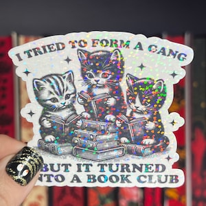 May include: A white sticker with a glittery background featuring three black and white kittens reading books. The sticker says "I tried to form a gang but it turned into a book club".