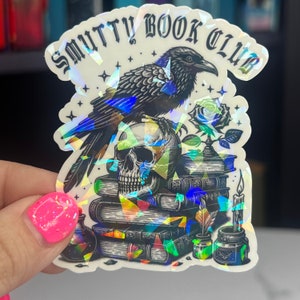 May include: A black raven perched on a stack of books with a skull and a rose. The sticker has a holographic finish and says "Smutty Book Club".