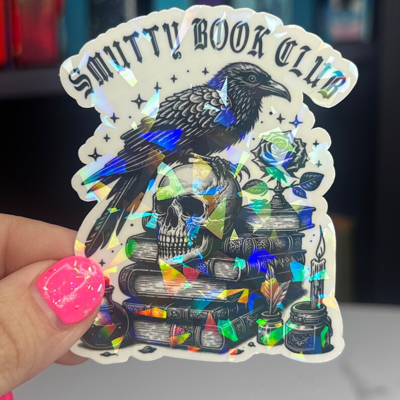 May include: A black raven perched on a stack of books with a skull and a rose. The sticker has a holographic finish and says "Smutty Book Club".