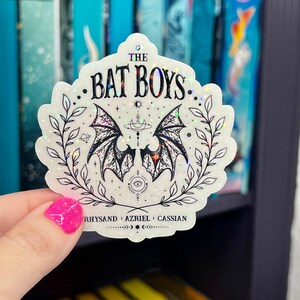 Pu&ograve; includere: Un adesivo bianco con finitura olografica che presenta un'illustrazione nera di ali di pipistrello con una luna crescente e stelle. Il testo "THE BAT BOYS" &egrave; scritto in nero sopra le ali. Sotto le ali, il testo "RHYSAND + AZRIEL + CASSIAN" &egrave; scritto in nero.