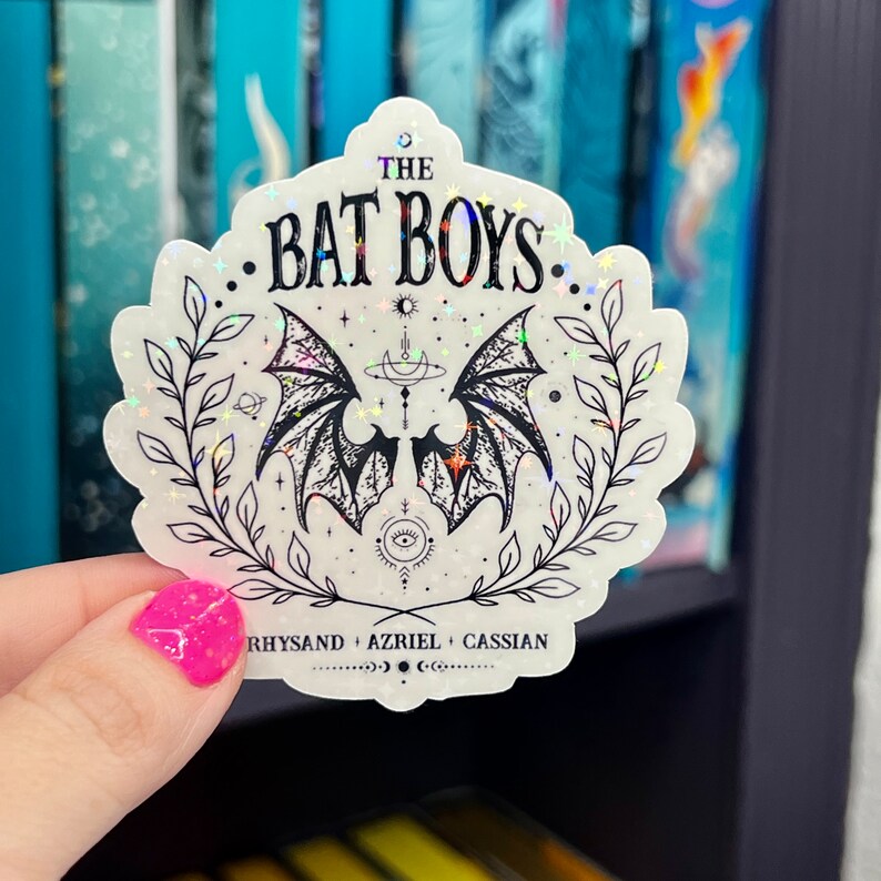 Pu&ograve; includere: Un adesivo bianco con finitura olografica che presenta un'illustrazione nera di ali di pipistrello con una luna crescente e stelle. Il testo "THE BAT BOYS" &egrave; scritto in nero sopra le ali. Sotto le ali, il testo "RHYSAND + AZRIEL + CASSIAN" &egrave; scritto in nero.