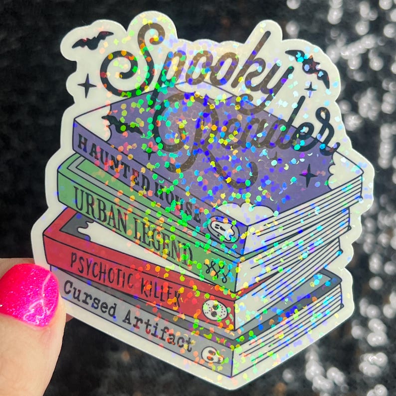 Op de afbeelding: Een sticker met een glinsterende, regenboogkleurige afwerking met een stapel boeken met de tekst "Spooky Reader" erop. De titels van de boeken zijn "Haunted House", "Urban Legend", "Psychotic Killer" en "Cursed Artifact".