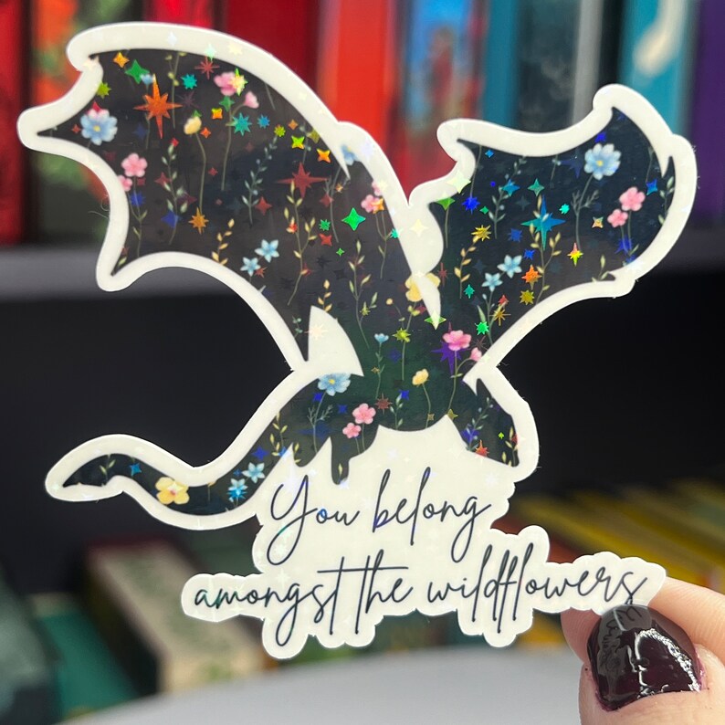 Op de afbeelding: Een zwarte holografische sticker met een bloemenpatroon en een draken silhouet. De sticker zegt "You belong amongst the wildflowers".