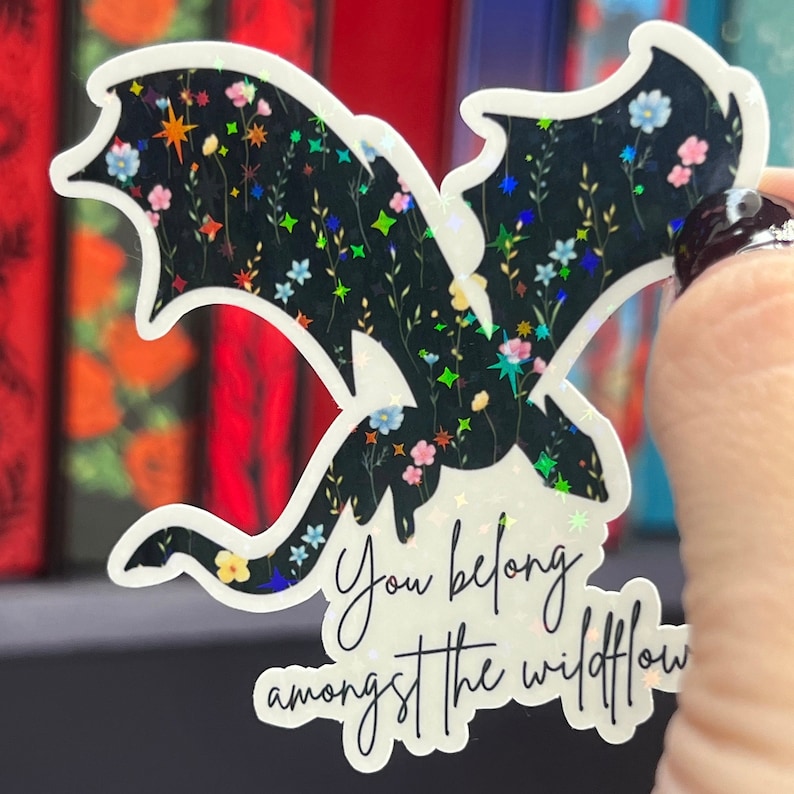 Op de afbeelding: Een transparante sticker met een zwart silhouet van een draak met bloemmotieven. De draak vliegt met zijn vleugels uitgespreid. De tekst "You belong amongst the wildflow" staat onder de draak.