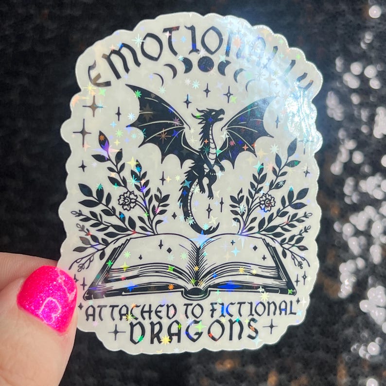 May include: A holographic sticker with a black and white illustration of a dragon emerging from an open book. The text reads "Emotionally attached to fictional dragons".