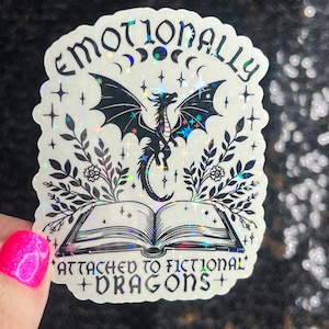 May include: A holographic sticker with a black dragon flying above an open book. The sticker has a white background with black text that reads "Emotionally Attached to Fictional Dragons".