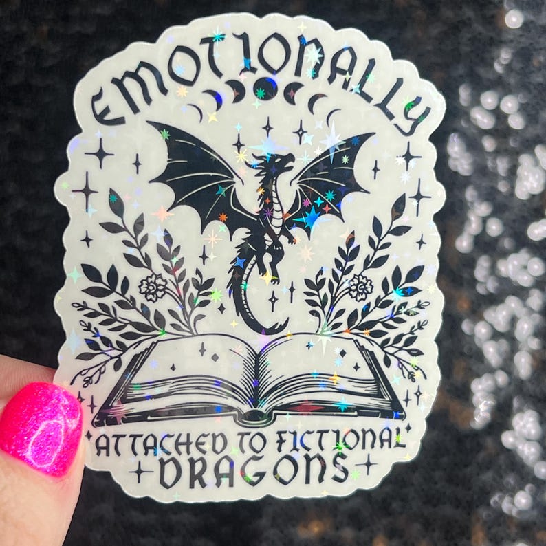 May include: A holographic sticker with a black dragon flying above an open book. The sticker has a white background with black text that reads "Emotionally Attached to Fictional Dragons".