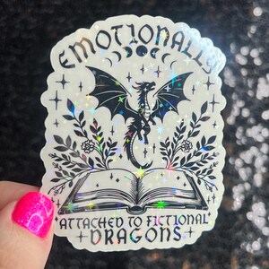 May include: A holographic sticker with a black and white illustration of a dragon emerging from an open book. The sticker has the text "Emotionally Attached to Fictional Dragons" around the top and bottom.