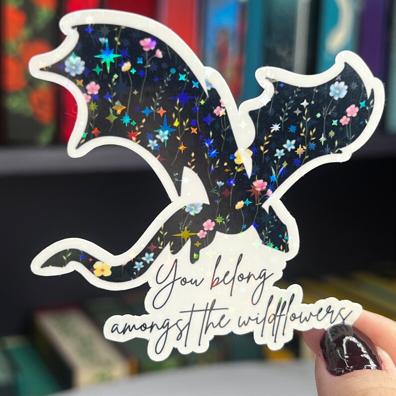 Op de afbeelding: Een zwarte holografische sticker met een draken silhouet en een bloemenpatroon. De sticker heeft de tekst "You belong amongst the wildflowers" in witte cursieve letters.