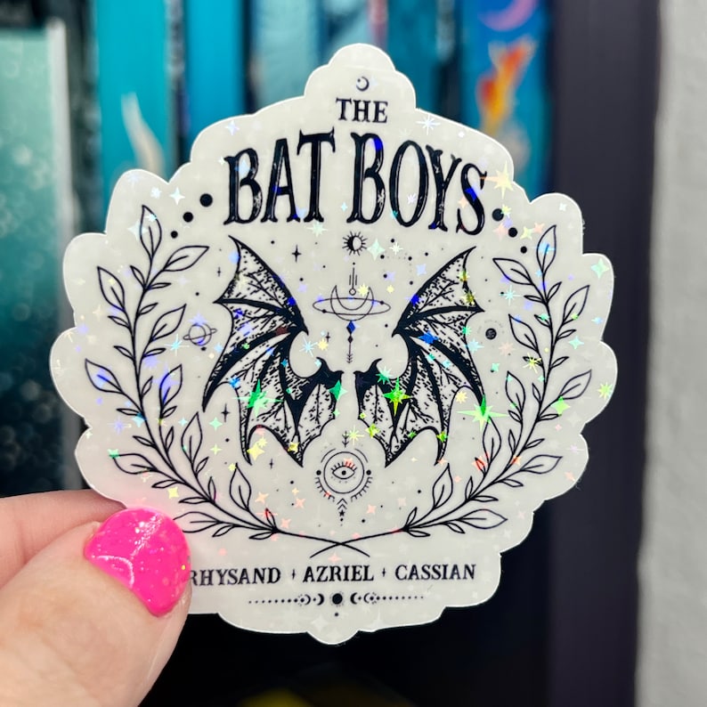 Pu&ograve; includere: Un adesivo bianco con finitura olografica che presenta un'illustrazione in bianco e nero di due ali di pipistrello con una luna crescente e stelle. Il testo "The Bat Boys" &egrave; scritto in lettere nere sopra le ali. Sotto le ali, il testo "Rhysand + Azriel + Cassian" &egrave; scritto in lettere nere.