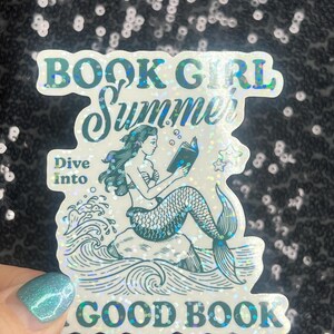 Może przedstawiać: Holograficzna naklejka z napisami "BOOK GIRL Summer" i "Dive Into GOOD BOOK". Naklejka przedstawia syrenę czytającą książkę siedzącą na skale, z bąbelkami i gwiazdami wok&oacute;ł niej.