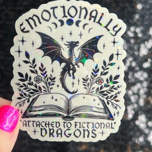 May include: A holographic sticker with a black and white illustration of a dragon flying over an open book. The text reads "Emotionally Attached to Fictional Dragons".