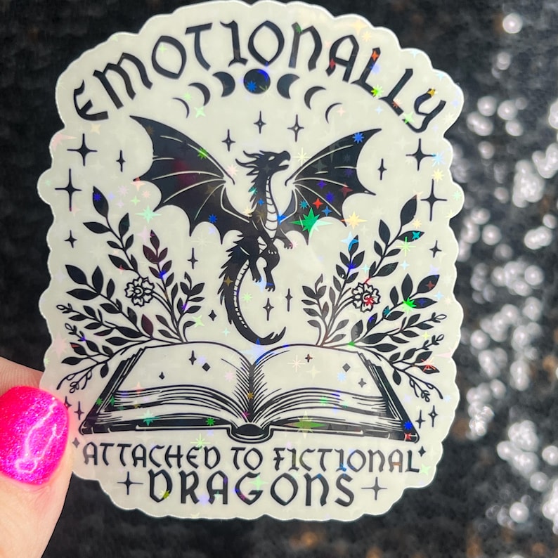 May include: A holographic sticker with a black and white illustration of a dragon flying over an open book. The text reads "Emotionally Attached to Fictional Dragons".