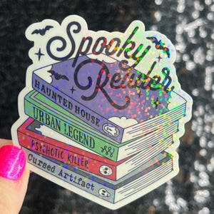 Op de afbeelding: Een holografische sticker met de tekst "Spooky Reader" en een stapel boeken met de titels "Haunted House", "Urban Legend", "Psychotic Killer" en "Cursed Artifact".