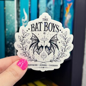 Pu&ograve; includere: Un adesivo bianco con un design nero di ali di pipistrello, stelle e una luna crescente. Il testo "The Bat Boys" &egrave; scritto in un font gotico. I nomi Rhysand, Azriel e Cassian sono elencati sotto le ali.