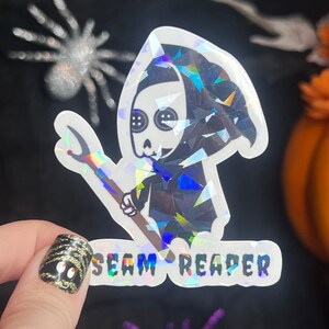 May include: A holographic sticker featuring a cartoon Grim Reaper holding a needle and thread. The sticker says "Seam Reaper" in a holographic font.