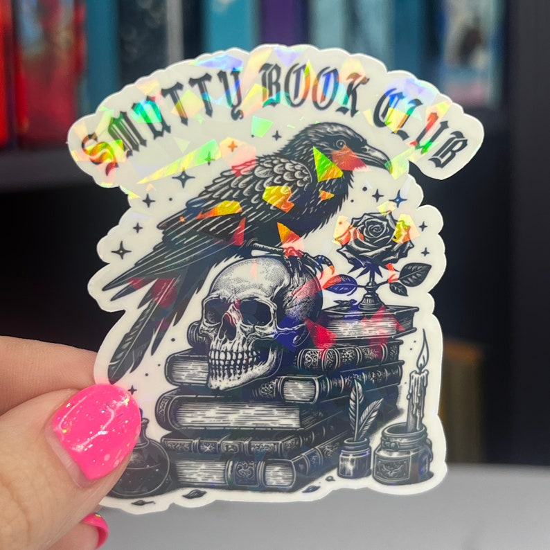 May include: A holographic sticker with a black raven perched on a stack of books. The raven is holding a skull in its talons. The sticker says "Smutty Book Club" in a gothic font.