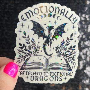 May include: A holographic sticker with a black dragon, a book, and floral accents. The text reads "Emotionally Attached to Fictional Dragons".
