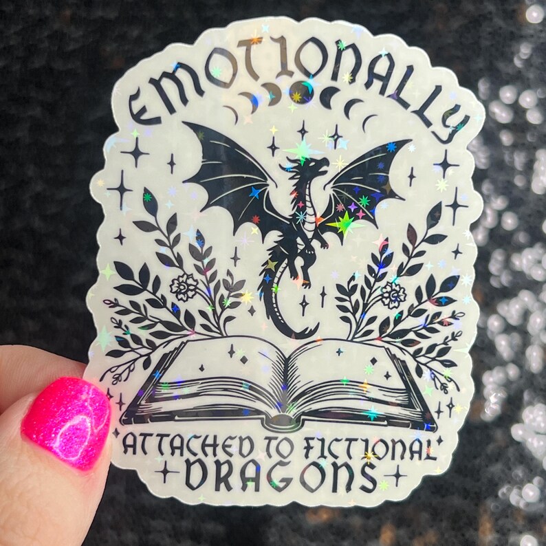 May include: A holographic sticker with a black dragon, a book, and floral accents. The text reads "Emotionally Attached to Fictional Dragons".