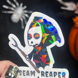 May include: A holographic sticker featuring a cartoon Grim Reaper holding a scythe. The reaper is wearing a black hooded robe with a skull face and large eyes. The text "SEAM REAPER" is printed below the reaper.