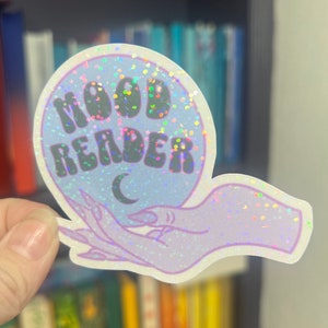 May include: A holographic sticker with a blue background and glitter. The sticker features a hand holding a crystal ball with the text "MOOD READER" and a crescent moon inside.