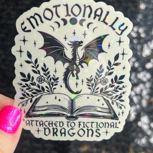 May include: A holographic sticker with a black and white illustration of a dragon, a book, and floral elements. The text reads "Emotionally Attached to Fictional Dragons".
