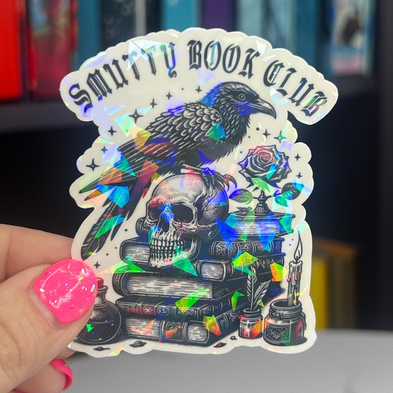 May include: A holographic sticker with a black raven perched on a skull, surrounded by a stack of books, a rose, and a candle. The sticker reads "Smutty Book Club".
