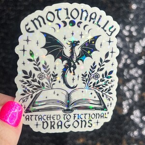 May include: A holographic sticker with a black dragon, a book, and the text "Emotionally Attached to Fictional Dragons".