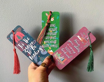 May include: Three Christmas-themed bookmarks with festive designs and tassels. The bookmarks feature illustrations of a Christmas tree, a snowman, and a stack of books. The text on the bookmarks reads "Tis the season to be reading", "Have your shelf a merry little bookmas", and "Books, chocolate, gingerbread, fairy lights, movie nights, festive jumpers".