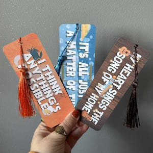 May include: Three rectangular bookmarks with tassels. One is orange with the text "I THINK ANYTHING'S POSSIBLE" and a crab illustration. Another is blue with the text "IT'S ALL JUST A MATTER OF T". The third is brown with the text "MY HEART SINGS THE SONG OF MY HOME".