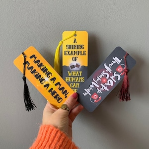 May include: Three bookmarks with different designs. The first bookmark is yellow with black text that says "Making a man, making a hero". The second bookmark is yellow with black text that says "A shining example of what humans can be". The third bookmark is black with red text that says "Salonpus Through Stormy Weather".