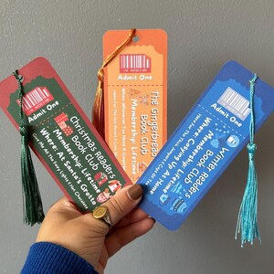 Può includere: Tre segnalibri colorati con nappe, di colore rosso, arancione e blu. Ogni segnalibro presenta la scritta "Admit One" e un tema diverso: Natale, Pan di Zenzero e Club di lettura invernale. Sfondo grigio.