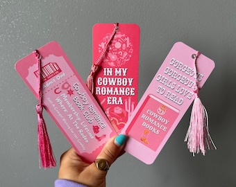 May include: Three pink bookmarks with pink tassels. The bookmarks feature cowboy-themed illustrations and text, including "In My Cowboy Romance Era", "Cowboy Romance Book Club", and "Gorgeous Girls Love to Read".
