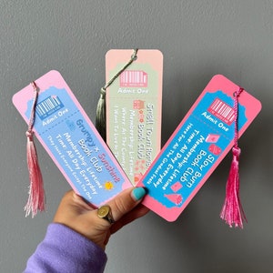 May include: Three pink and blue ticket-style bookmarks with colourful illustrations and text. The bookmarks have tassels. The text on the bookmarks reads "Grumpy x Sunshine Book Club", "Slow Burn Book Club", and "Small Town Roma Book Club".