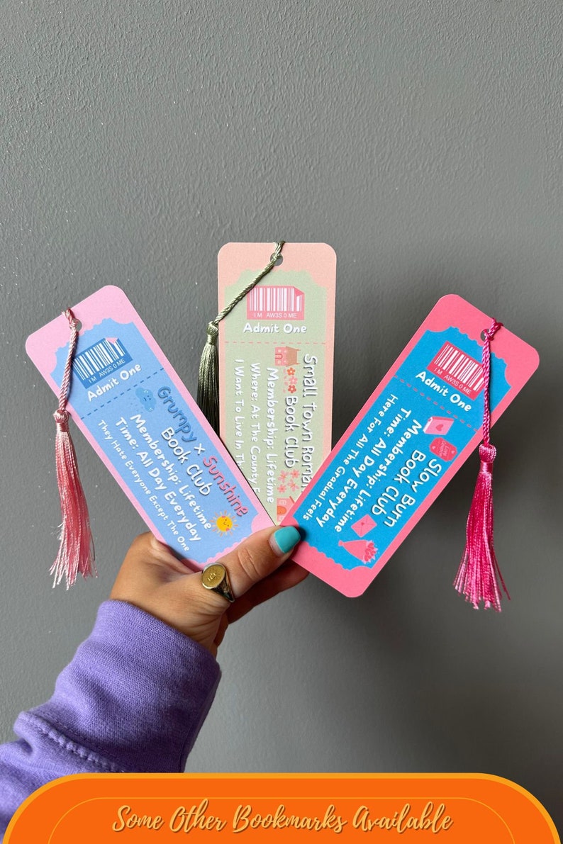 May include: Three pink and blue ticket-style bookmarks with colourful illustrations and text. The bookmarks have tassels. The text on the bookmarks reads "Grumpy x Sunshine Book Club", "Slow Burn Book Club", and "Small Town Roma Book Club".