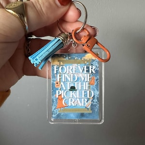 以下が含まれることがあります： 「FOREVER FIND ME AT THE PICKLED CRAB」の白文字が入った、透明な長方形のキーホルダー。キーホルダーには、青いタッセル、シルバーのキーリング、オレンジ色の留め金が付いています。
