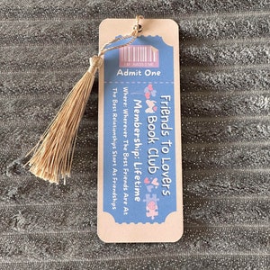 May include: A light blue and pink bookmark with a ticket design. It says "Friends to Lovers Book Club" and "Membership: Lifetime".