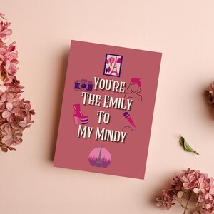 May include: A mauve greeting card with the text "You're the Emily to my Mindy" and illustrations of a camera, shoe, beret, microphone, and Eiffel Tower. Pink flowers are arranged around the card on a light pink surface.