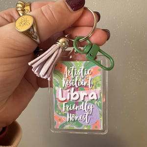 Könnte beinhalten: Ein klarer, rechteckiger Schlüsselanhänger mit farbenfrohem Design und dem Wort "Libra" in Weiß. Der Schlüsselanhänger enthält auch die Wörter "Artistic, Resilient, Friendly, Honest". Er hat eine hellviolette Quaste und einen grünen Verschluss.